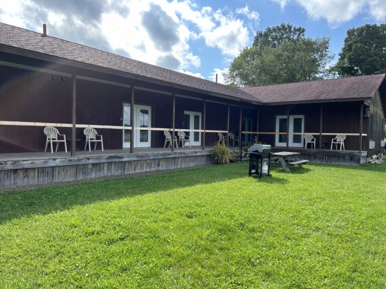 A wooden building with a porch, white chairs, picnic table, and a grill on a grassy lawn under a partly cloudy sky.